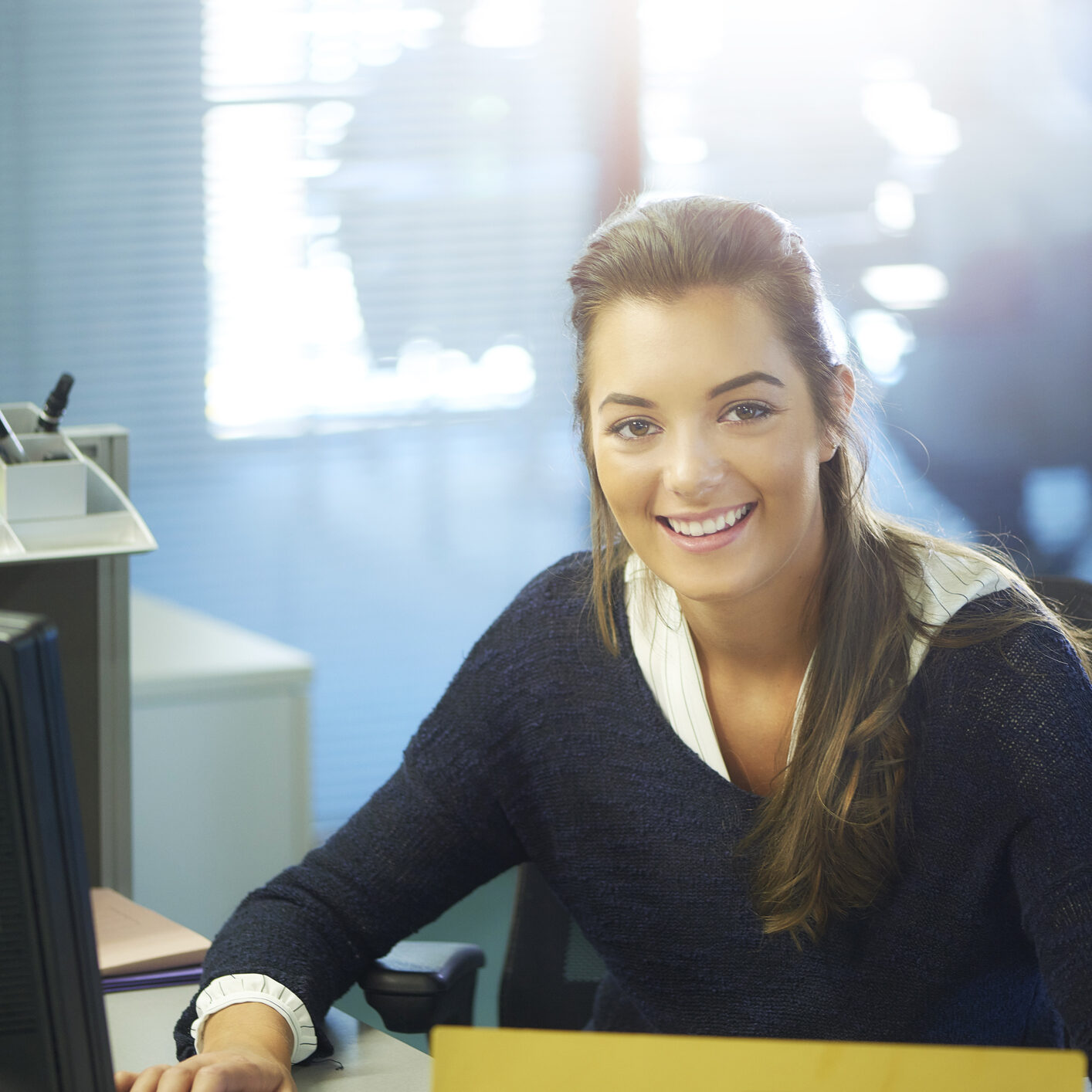 young female office worker or work experience student , sitting at her desk and looking at the camera . She is smiling to camera and looks very happy in her work.